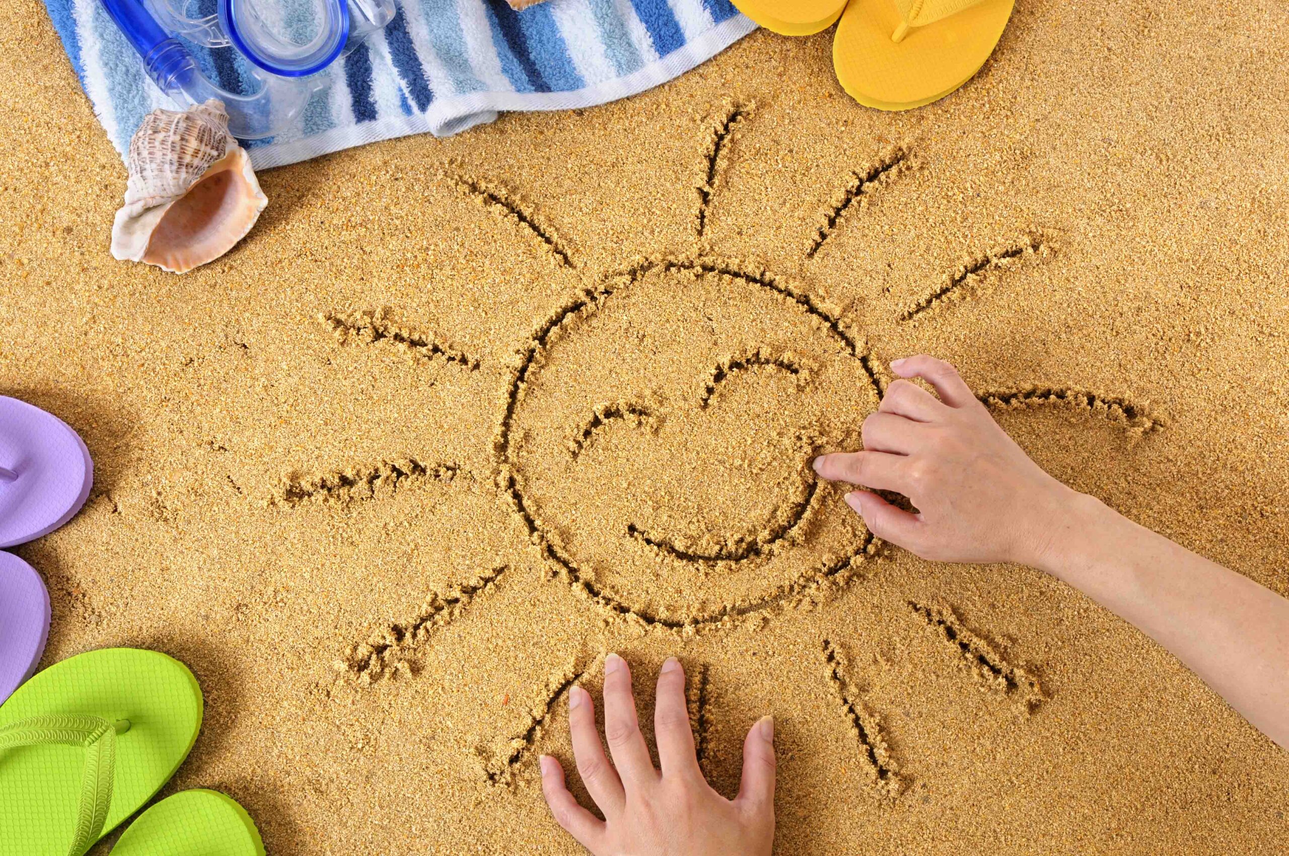 Child drawing a smiley sun in sand with towel, seashells and flip flops